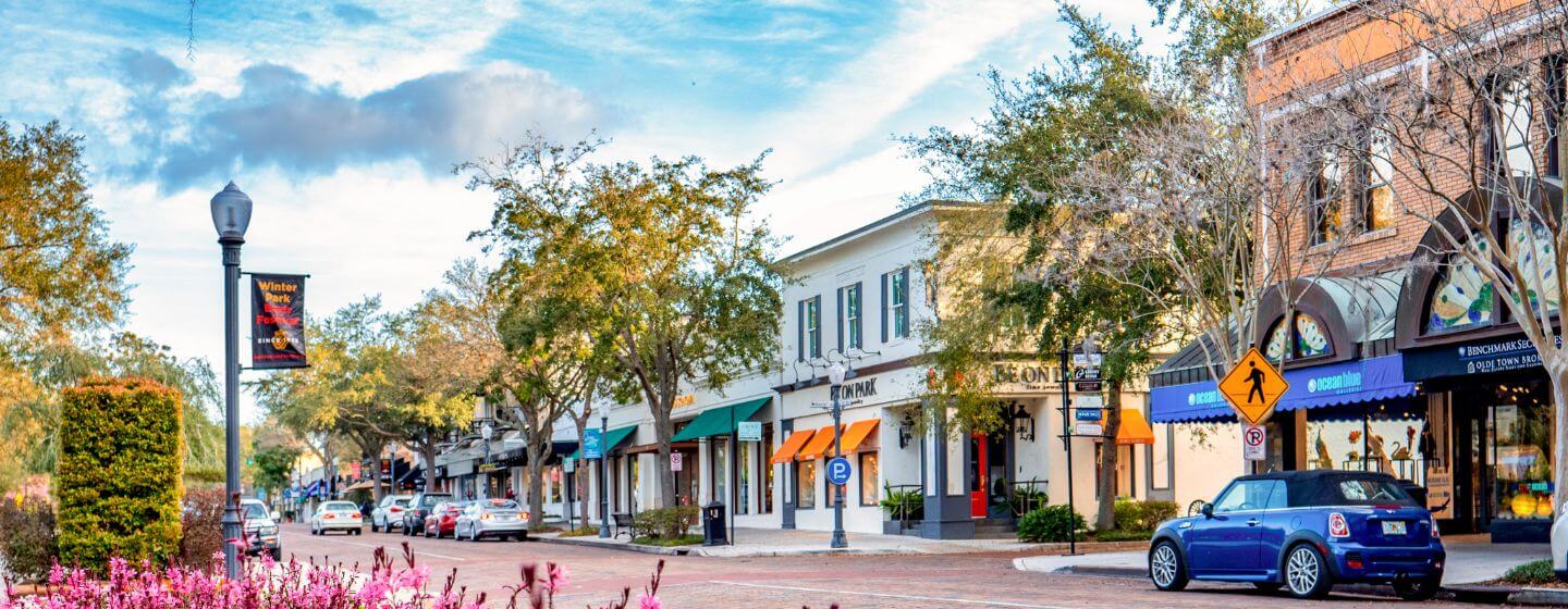 A tree-lined street with parked cars, storefronts with awnings, a brick sidewalk, and a crosswalk invites you to just move it under a blue sky with clouds.