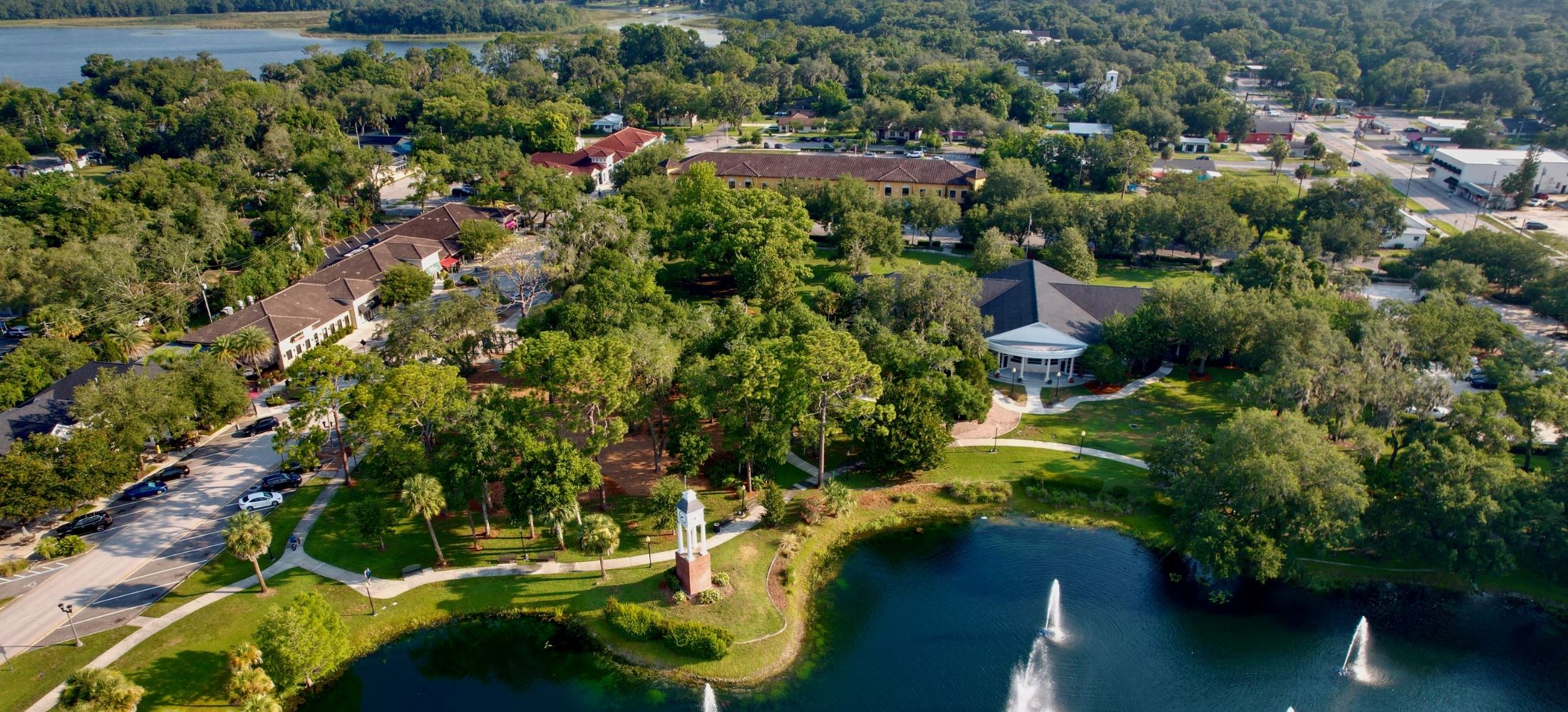 Aerial view of a small town with trees, buildings, a pond with fountains, and walking paths on a sunny day inviting you to just move it outdoors.