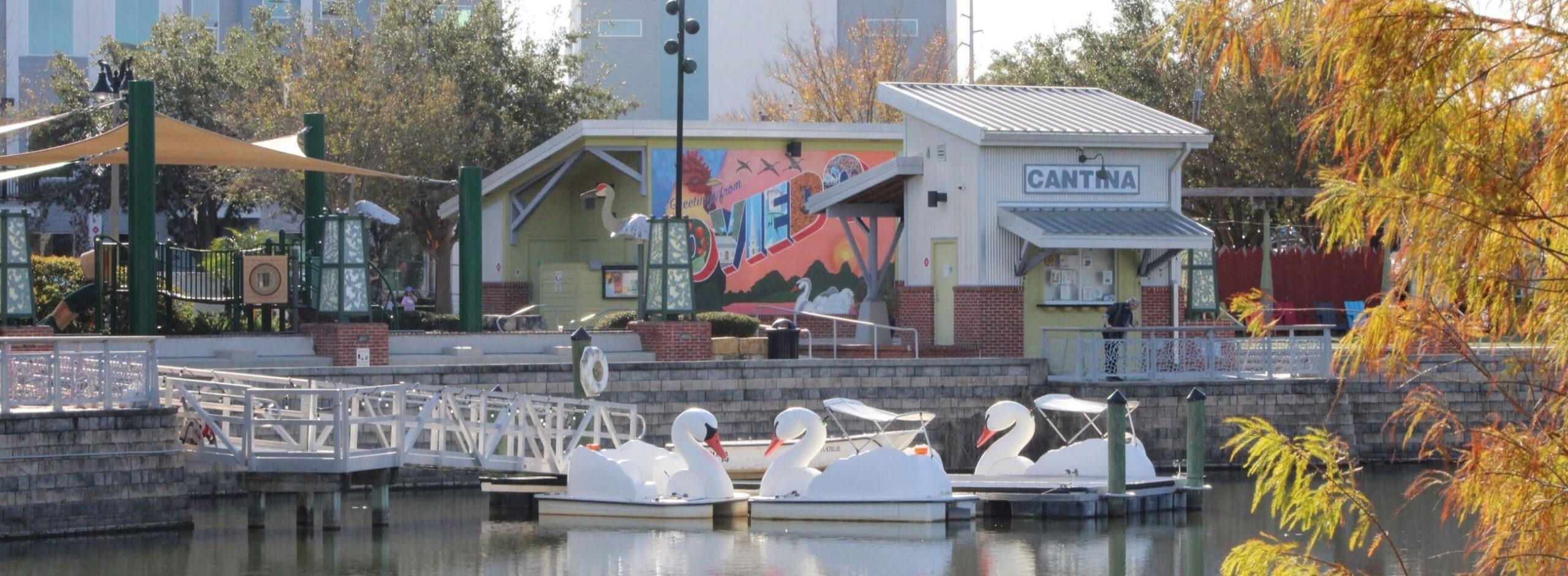 A small dock with swan-shaped pedal boats on a pond near a cantina building, featuring a mural and trees in the background—just move it for a fun lake adventure.